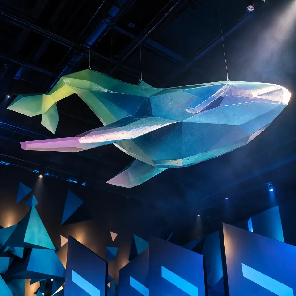 Ballena low-poly en tonos azules y verdes suspendida del techo del hub, sobre paneles y triángulos azules.
