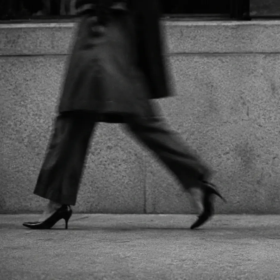 Fotografía en blanco y negro de dos personas caminando con prisa por una acera, junto a un muro de piedra.