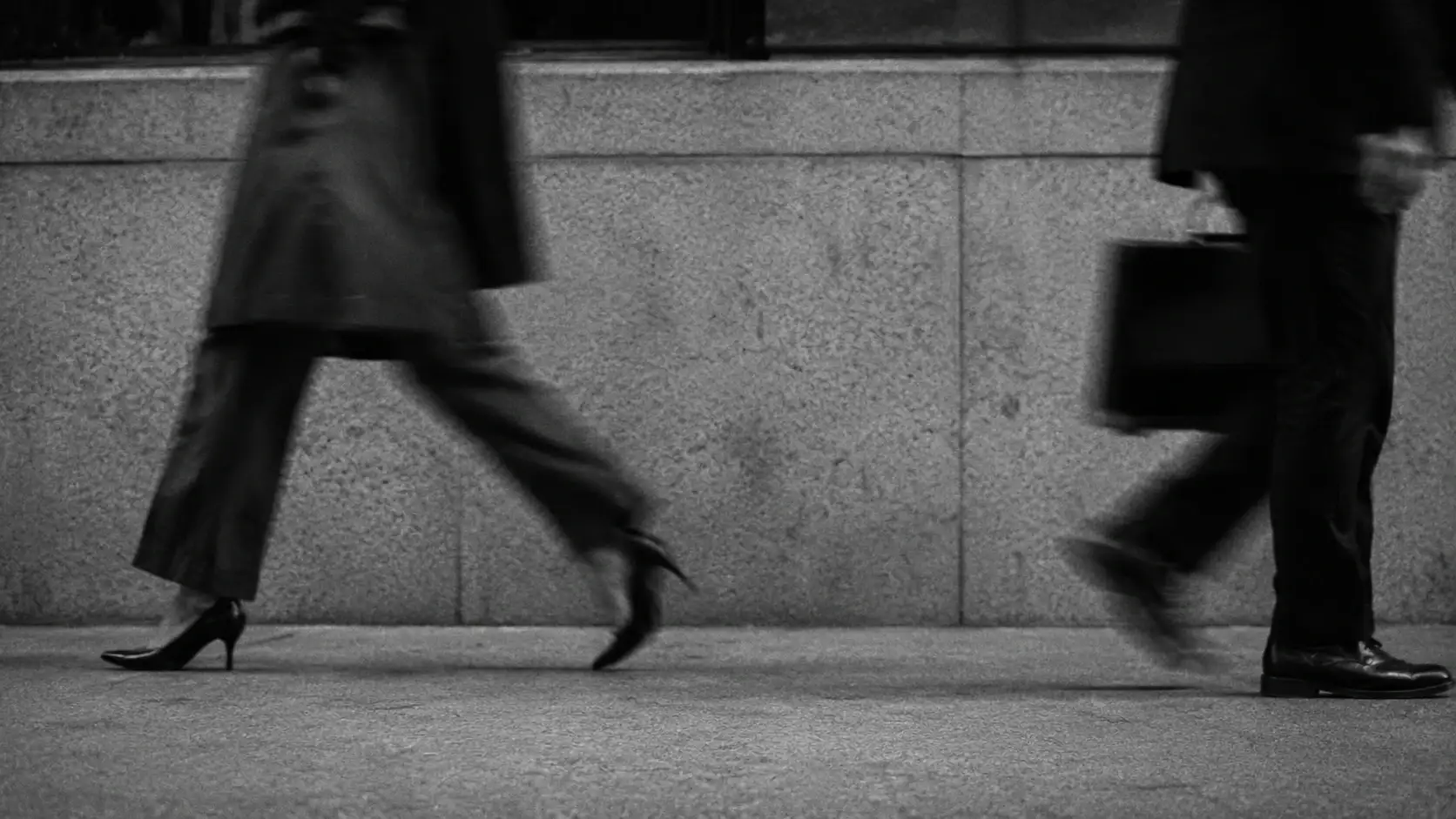 Fotografía en blanco y negro de dos personas caminando con prisa por una acera, junto a un muro de piedra.