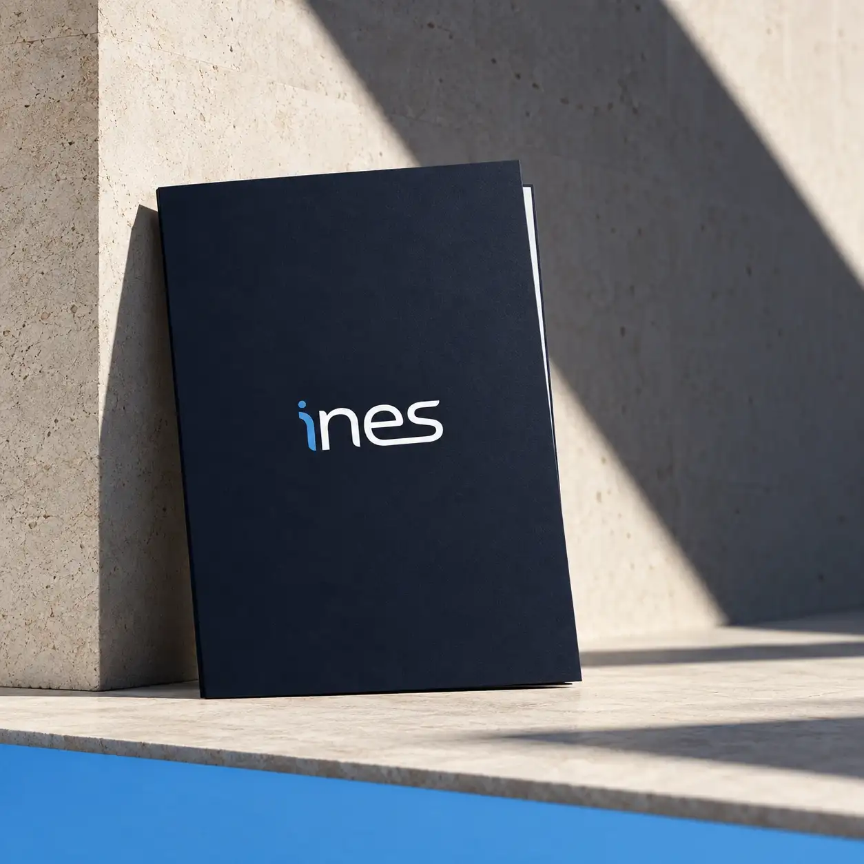 Carpeta corporativa azul noche con el logotipo de INES, apoyada en una pared de piedra.